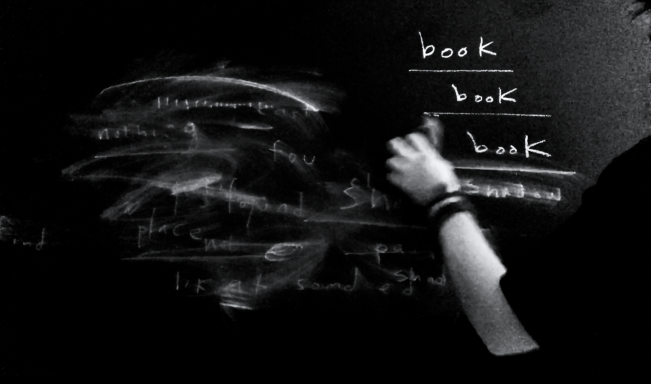 A black and white image of an arm writing on a black wall with white chalk. The text on the wall reads "book book book"